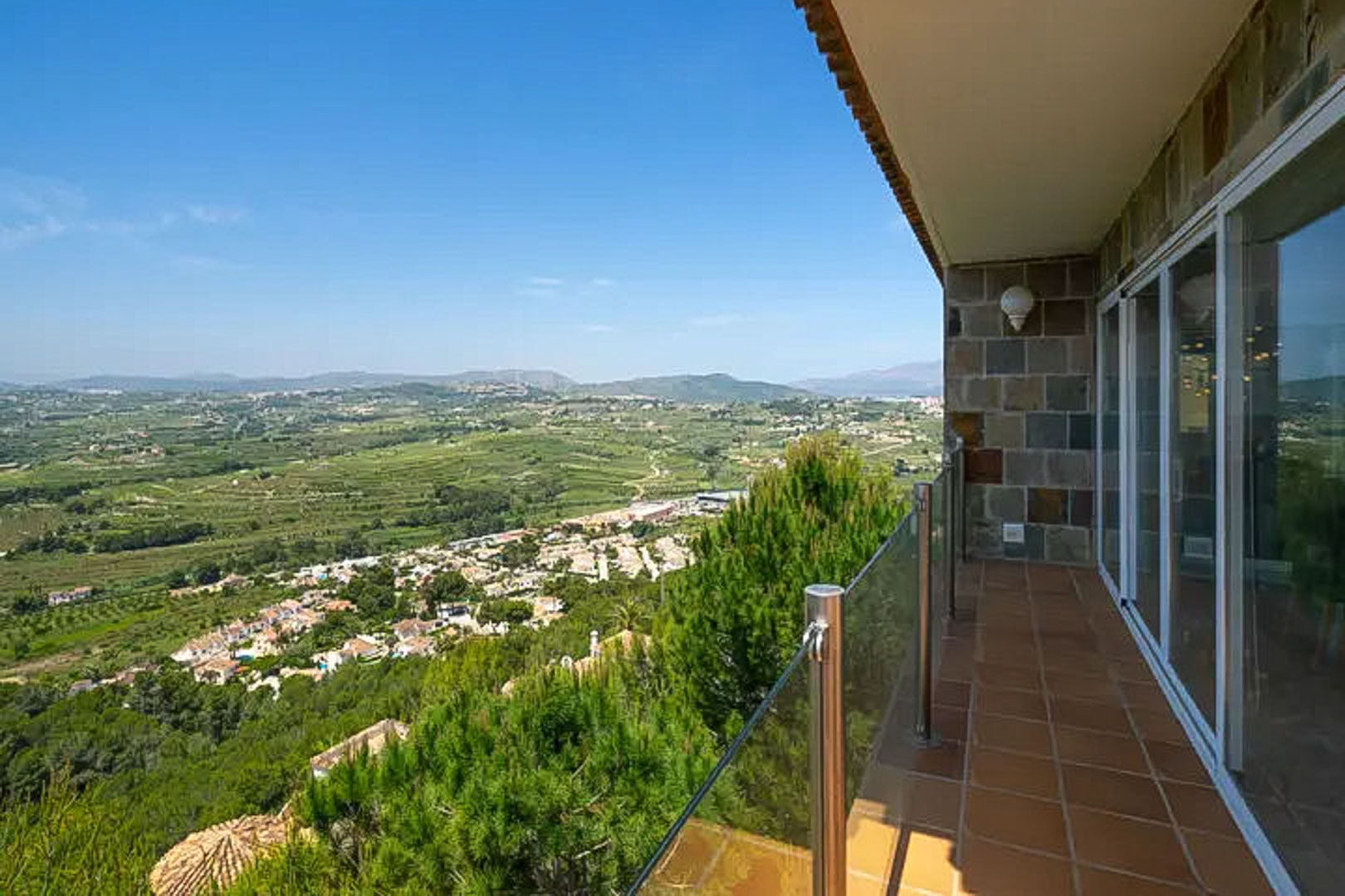 Terraza elevada con suelo de terracota y barandilla de cristal, ofreciendo vistas panorámicas al valle de Moraira y las montañas bajo un cielo despejado.