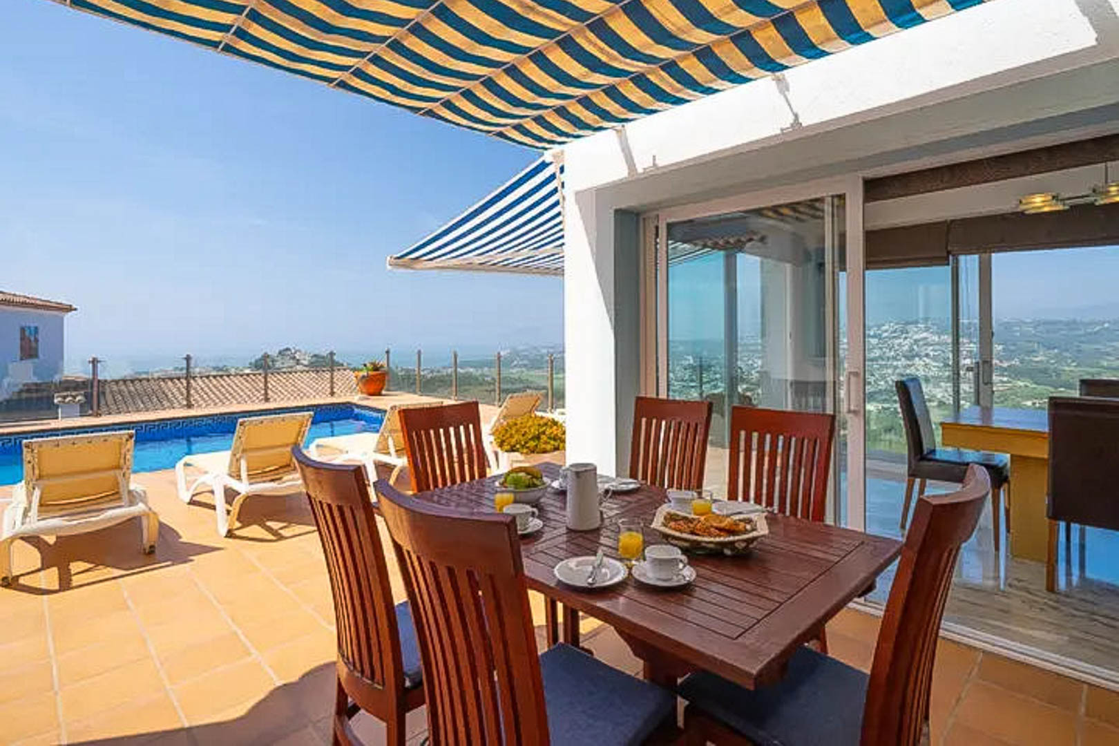 Terraza exterior con comedor de madera bajo un toldo de rayas, con vistas directas a la piscina privada y al litoral de Moraira.