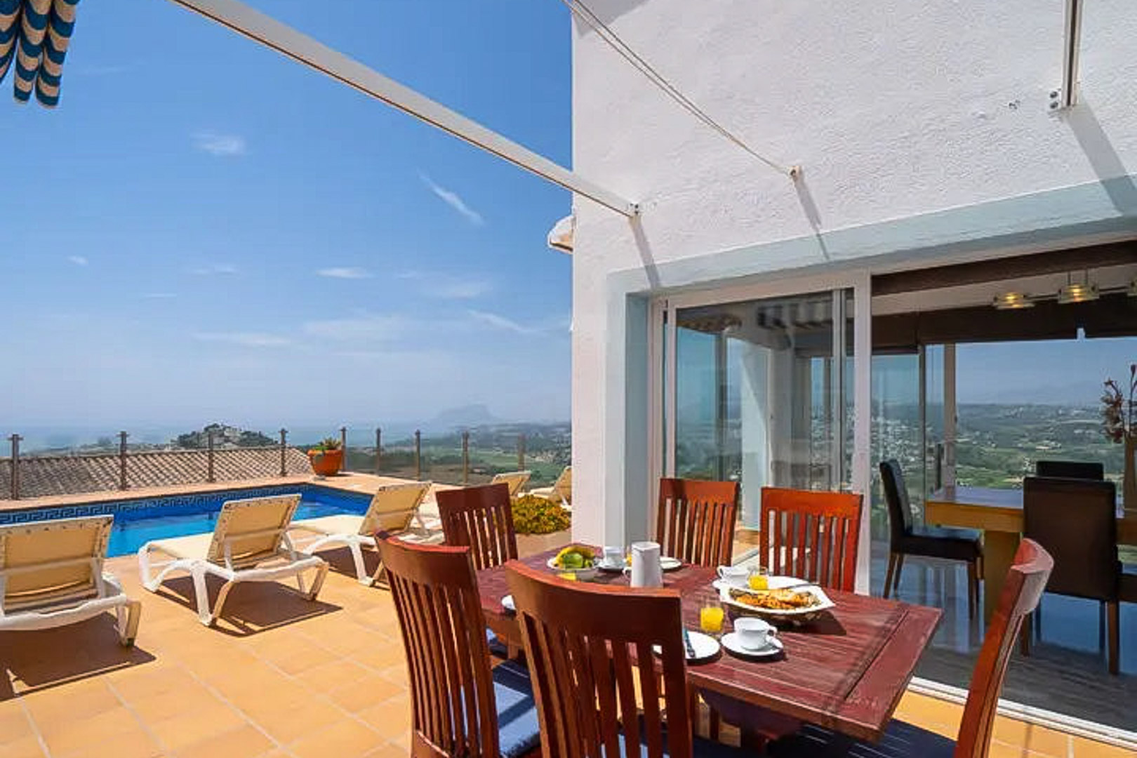 Terraza exterior con zona de comedor en madera, piscina privada con borde de mosaico y vistas panorámicas al mar y la costa de Moraira.