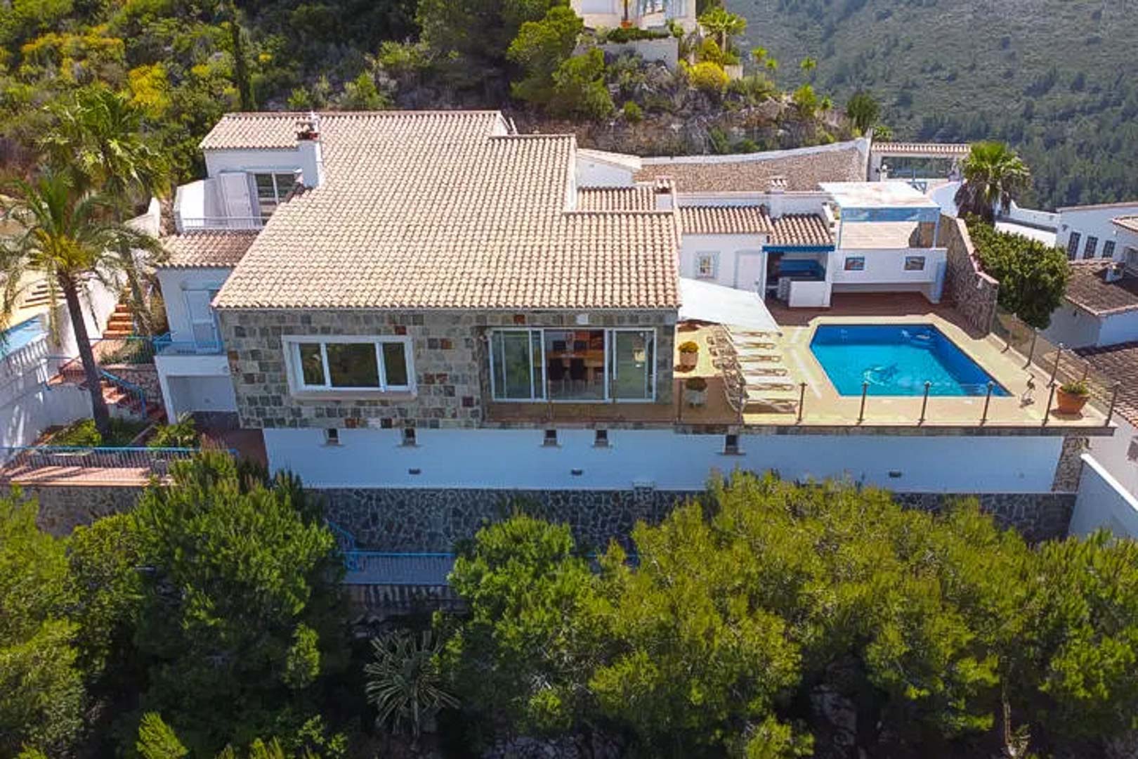 Vista aérea de la villa con fachada de piedra natural, tejados cerámicos y terraza principal con piscina privada rodeada de vegetación mediterránea.