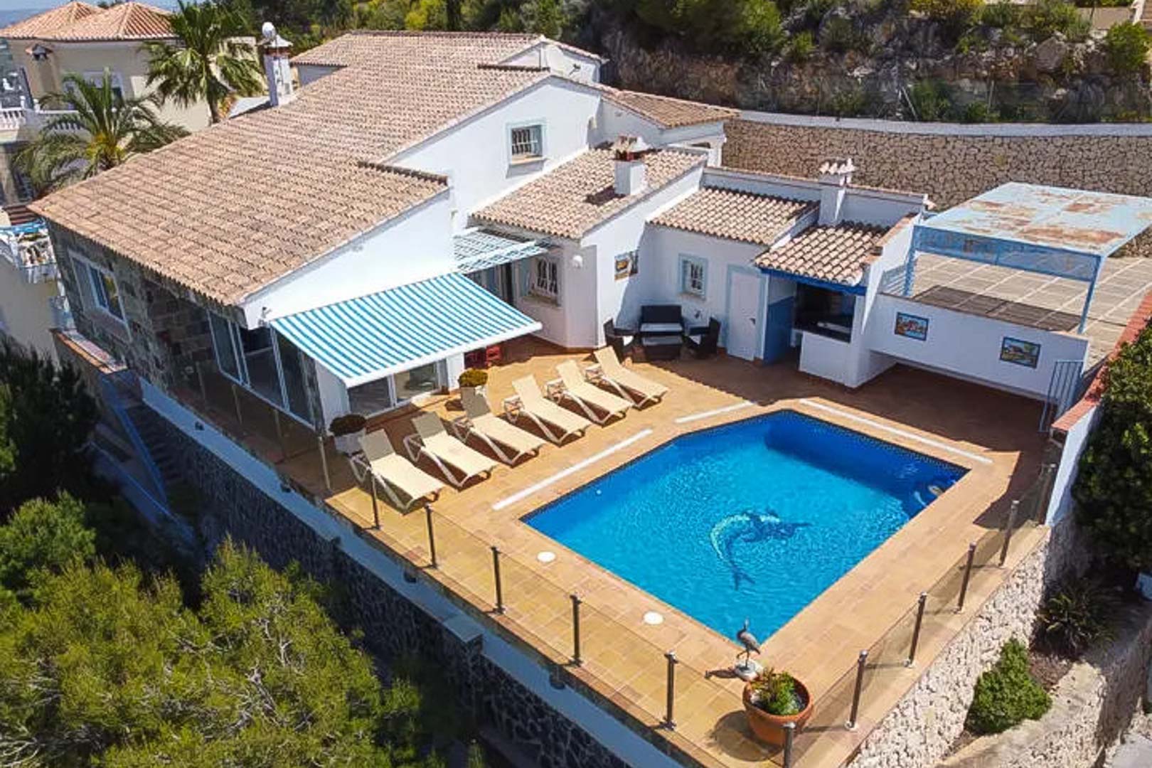 Vista aérea de la villa con piscina privada de mosaico, terraza con tumbonas, toldo de rayas y zona de bar exterior protegida por barandillas de cristal.