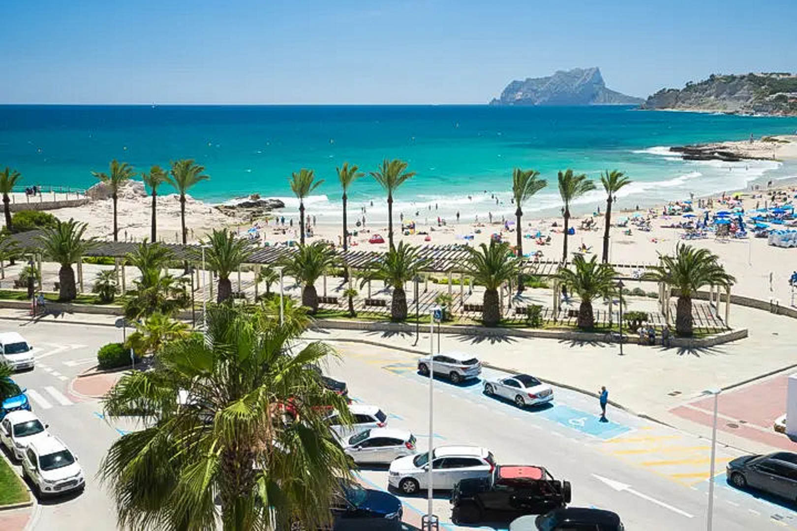 Vista panorámica de la playa de Moraira con aguas turquesas y el Peñón de Ifach al fondo, integrada en un entorno urbano con palmeras y paseo marítimo.