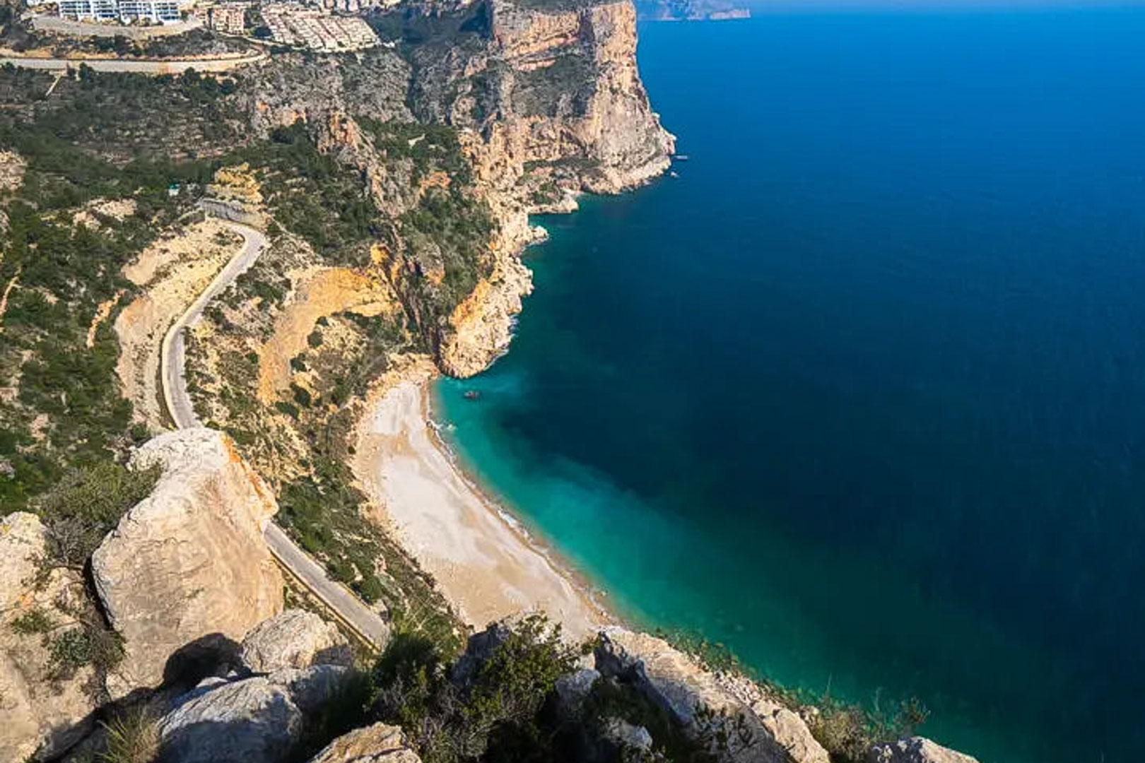 Vista panorámica de los acantilados de la Costa Blanca, con una cala de aguas turquesas y una carretera serpenteante que recorre el litoral mediterráneo.