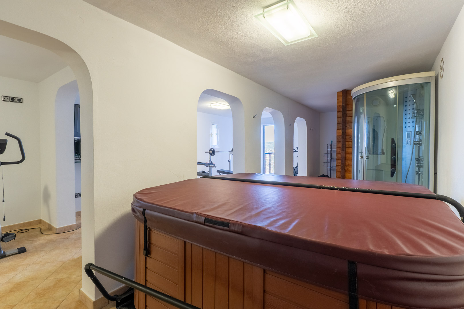 Zona de bienestar interior con jacuzzi cubierto, cabina de ducha con hidromasaje y acceso a través de arcos a un área de gimnasio equipada.