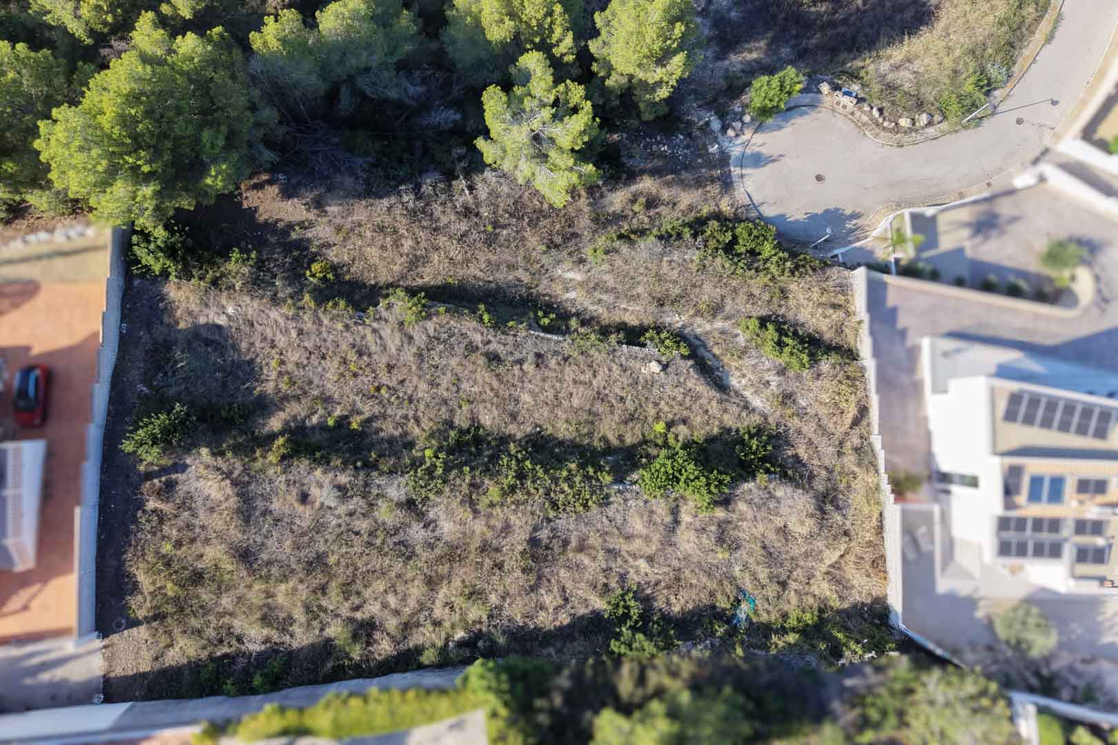 Vista aérea cenital de la parcela urbana escalonada con muros de piedra natural, rodeada de pinos y situada al final de una calle tranquila en Moraira.