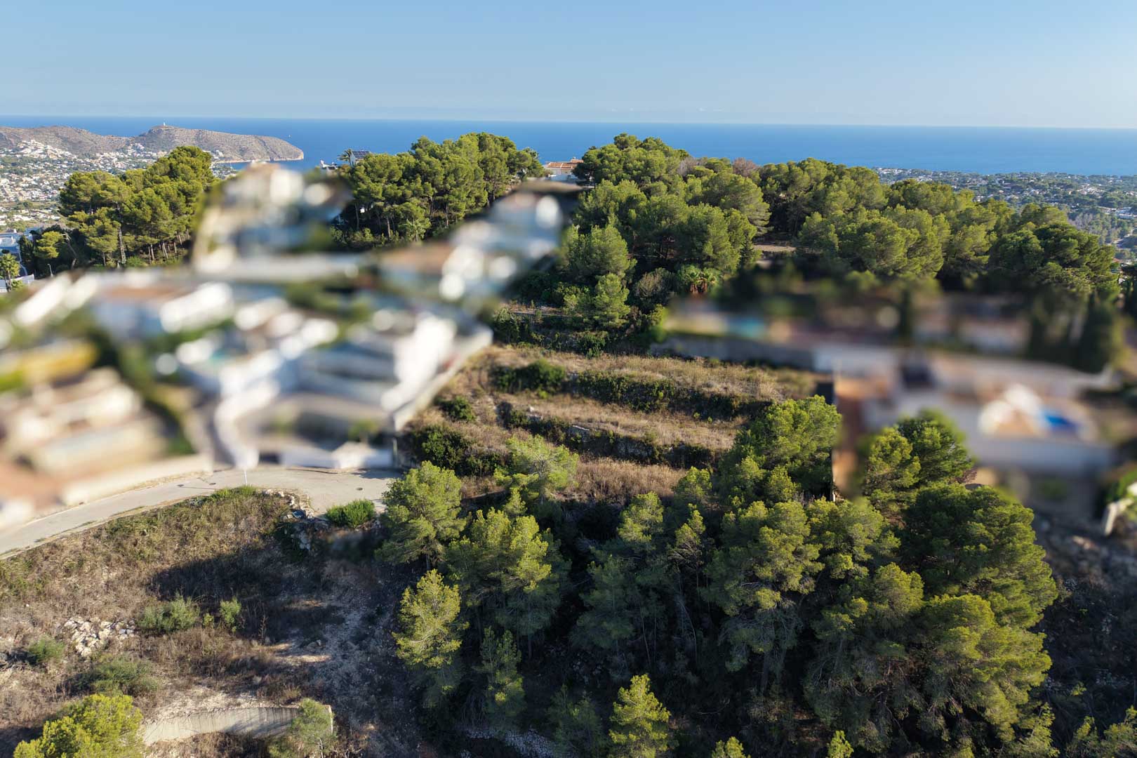 Vista aérea de la parcela urbana aterrazada, rodeada de densos pinos mediterráneos con vistas despejadas hacia el mar y el litoral de Moraira.