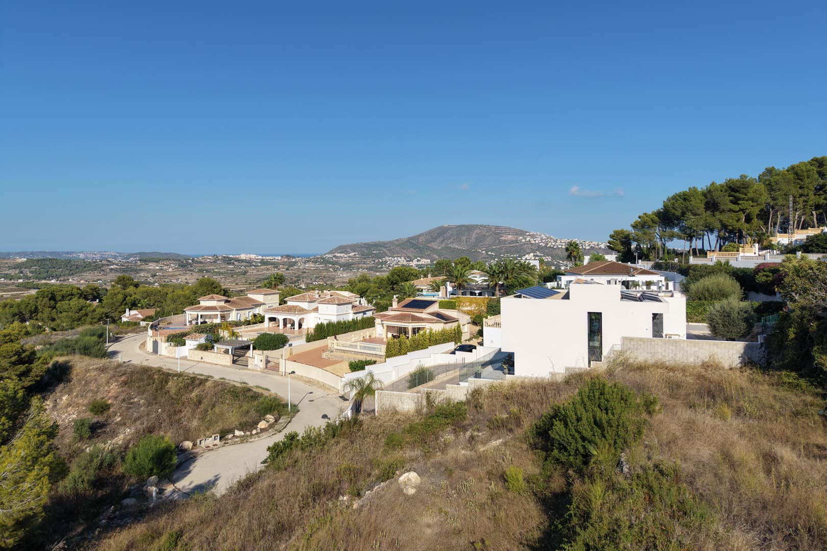 Vista elevada de la zona residencial con villas de estilo mediterráneo y moderno, rodeadas de pinos y con vistas abiertas hacia el valle y el mar.
