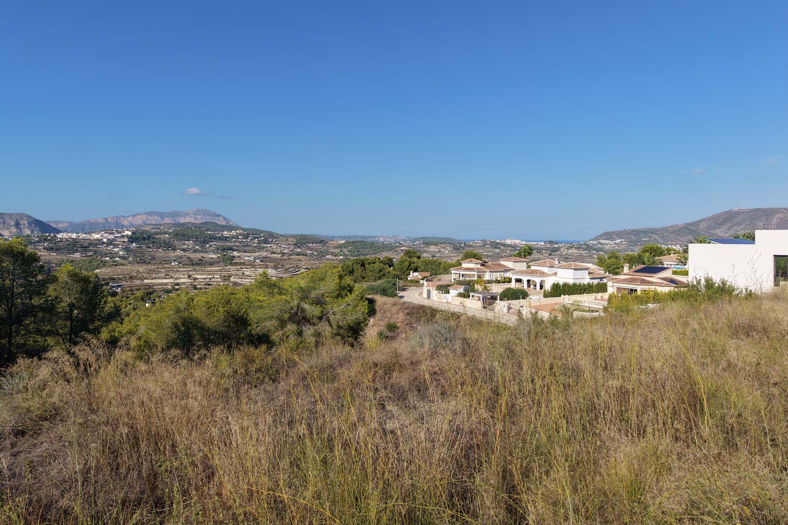 Parcela urbana elevada con vegetación natural, vistas despejadas al valle, montañas y destellos del mar Mediterráneo bajo un cielo despejado.