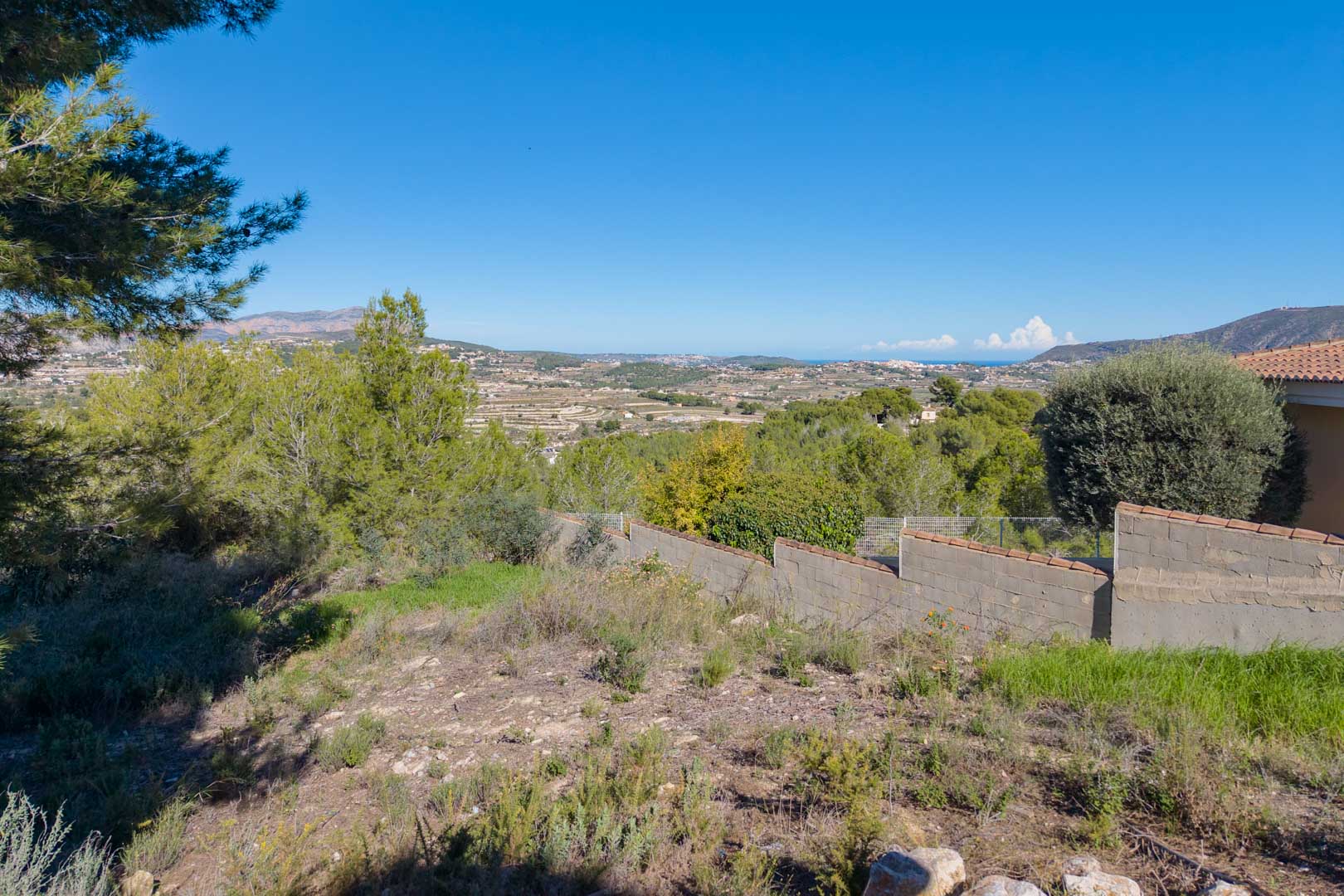 Parcela elevada con vistas panorámicas al valle y al mar. El terreno presenta vegetación autóctona y un muro perimetral de bloques con remate cerámico.