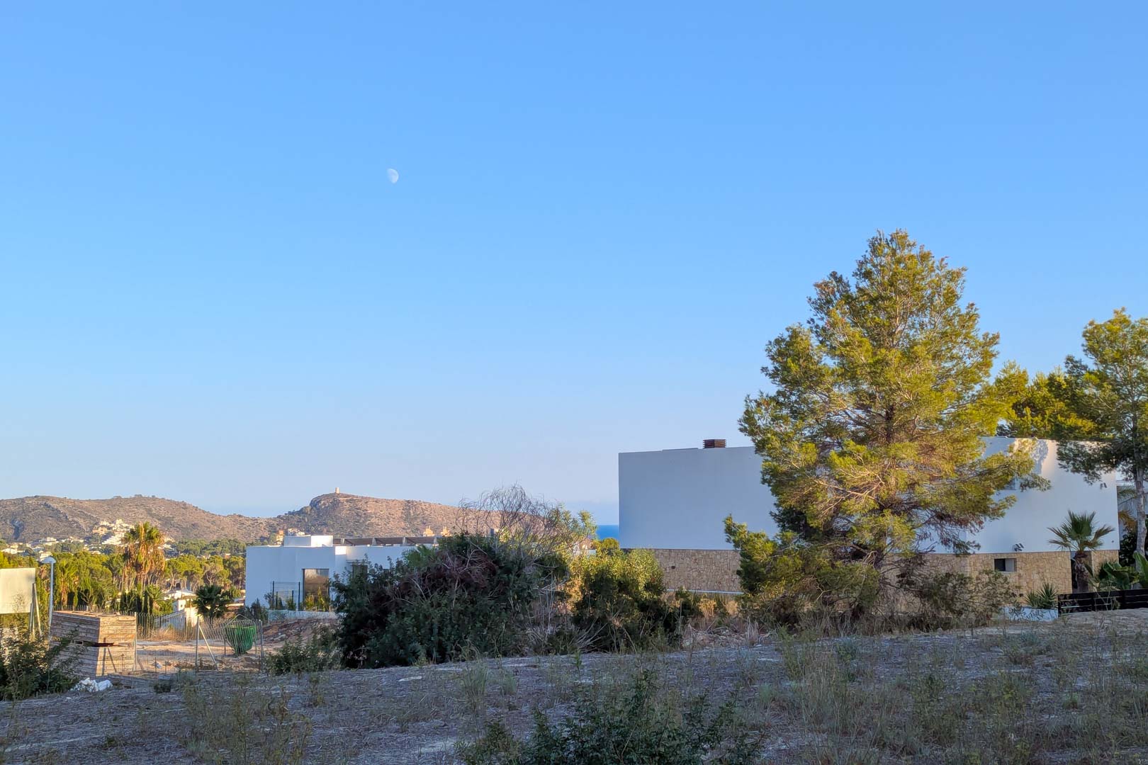 Vista desde la parcela hacia villas modernas de diseño cúbico, rodeadas de pinos mediterráneos con montañas y el mar en el horizonte.