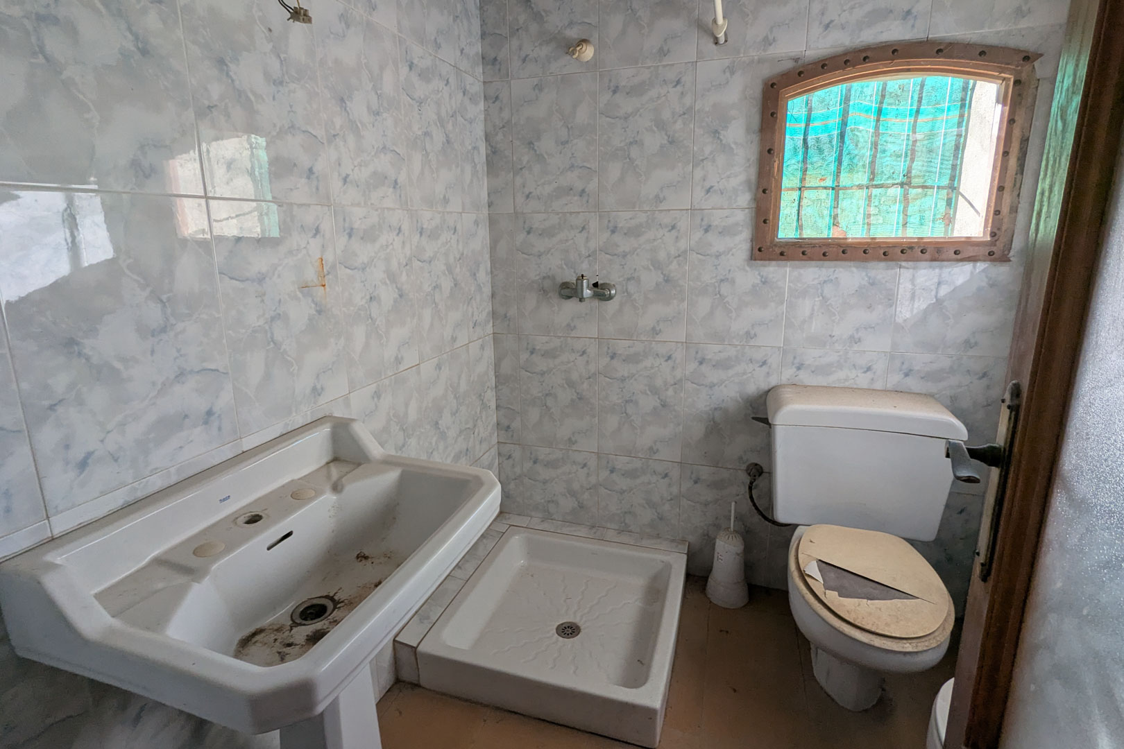 Cuarto de baño original con azulejos de efecto mármol azulado, lavabo de pedestal, plato de ducha cuadrado y una pequeña ventana de arco con marco de madera.