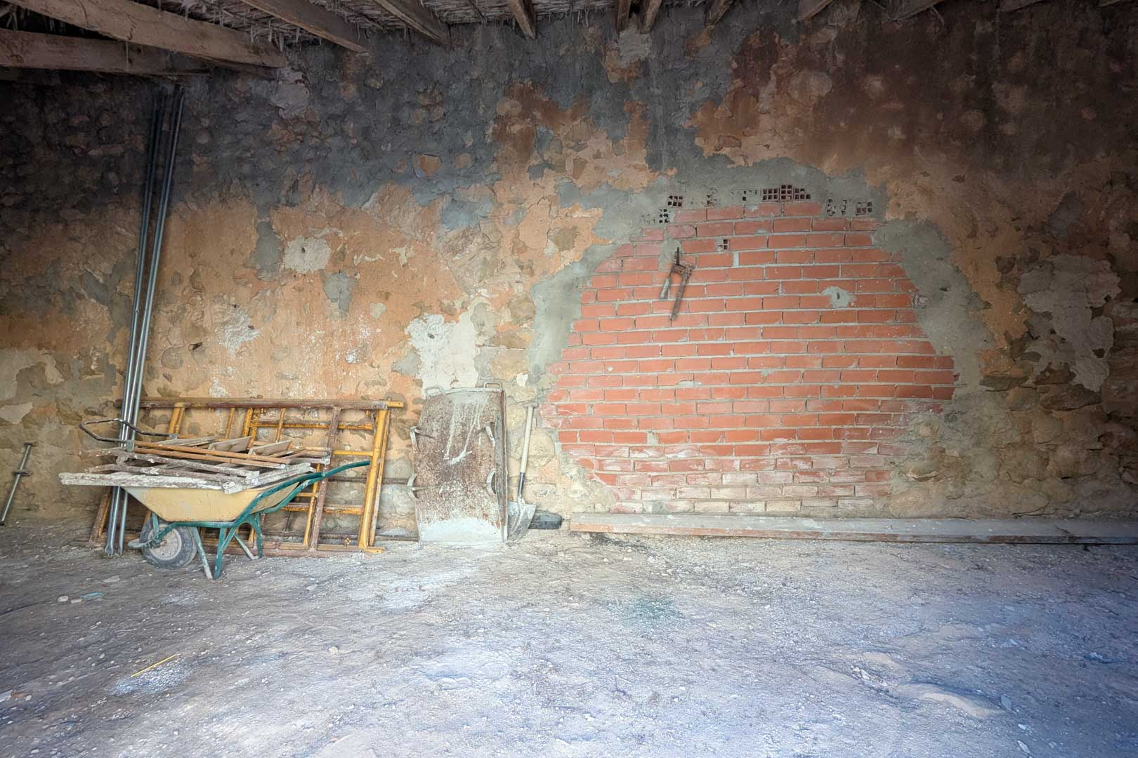 Espacio interior rústico con techos de vigas de madera y cañizo, paredes de piedra original y una sección de ladrillo visto. Incluye herramientas de obra y suelo de tierra.