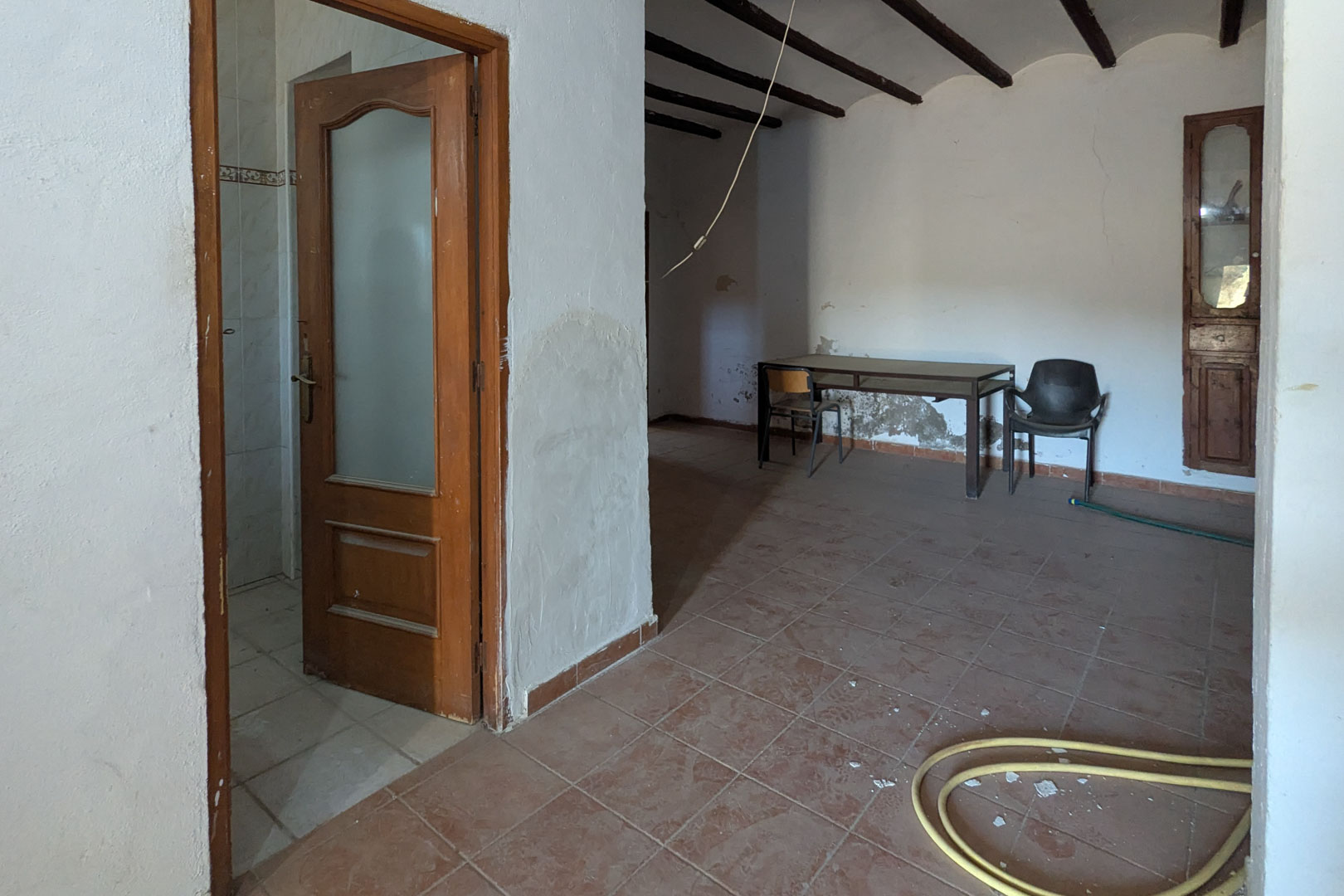 Interior rústico con vigas de madera a la vista, suelos de gres y paredes de cal. Incluye una puerta de madera con cristal y una alacena empotrada tradicional.