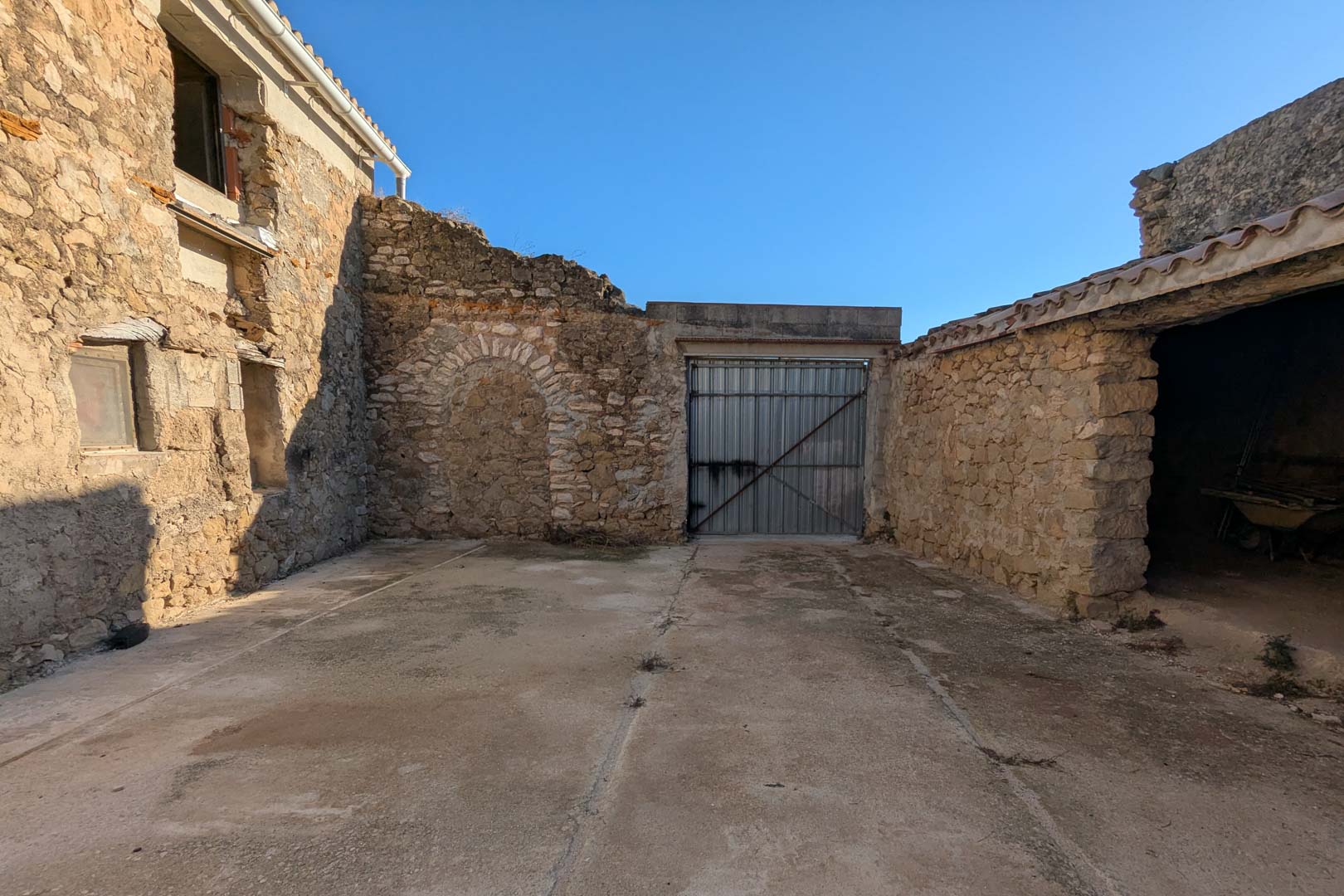 Patio exterior con muros de piedra natural, un arco original tapiado y acceso lateral a los antiguos establos de la finca.
