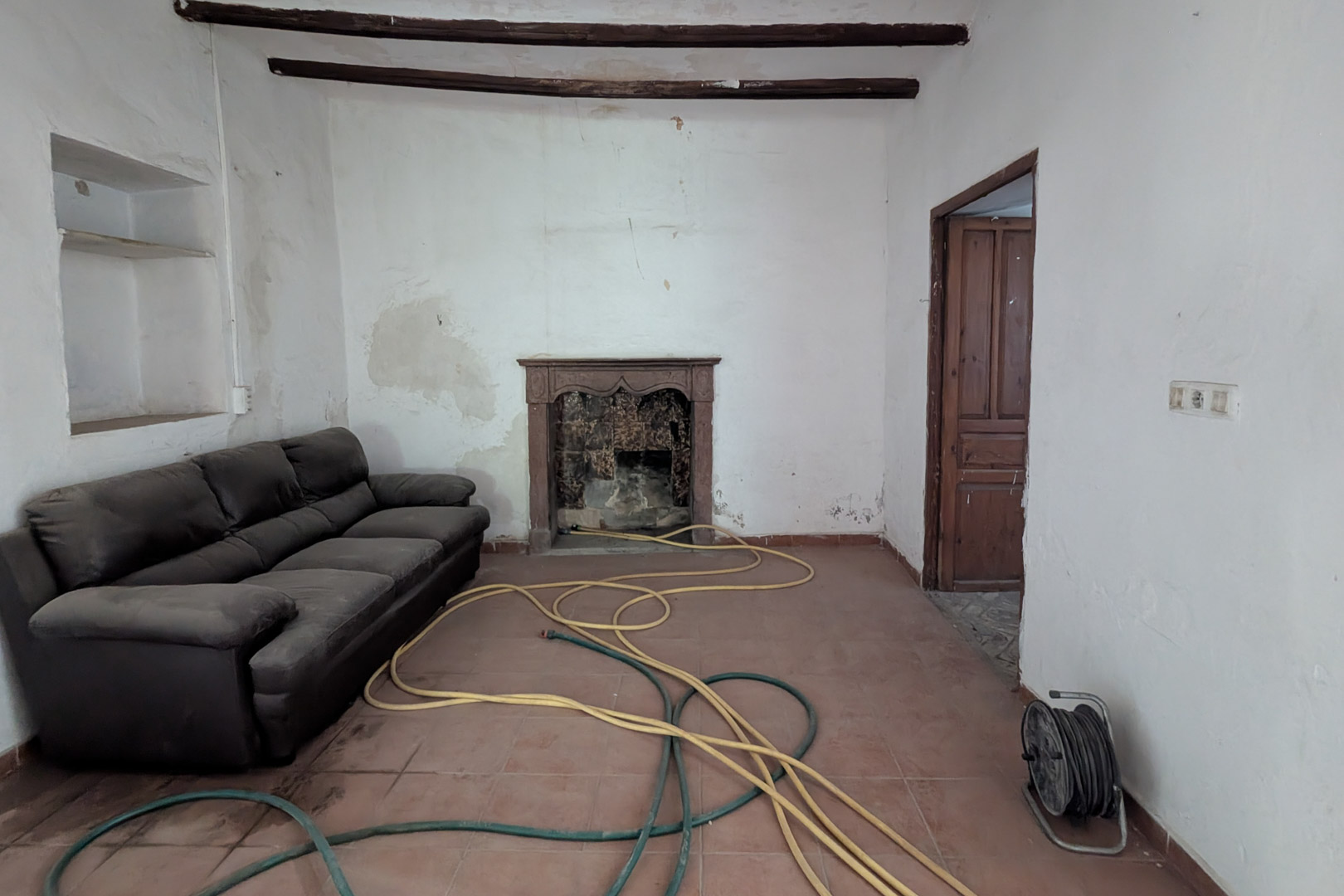 Salón tradicional con vigas de madera vista, suelo de baldosas de barro y una chimenea original con marco de piedra tallada. Las paredes conservan hornacinas integradas.