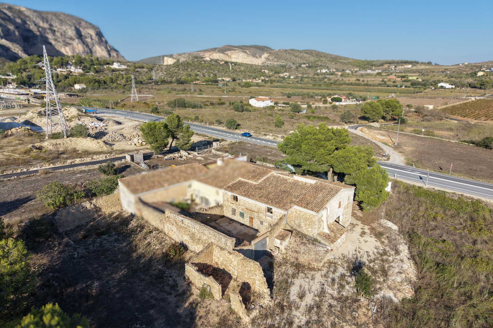 Vista aérea de la casa de campo tradicional con muros de piedra natural, tejados de teja cerámica y una parcela llana rodeada de montañas en Teulada.