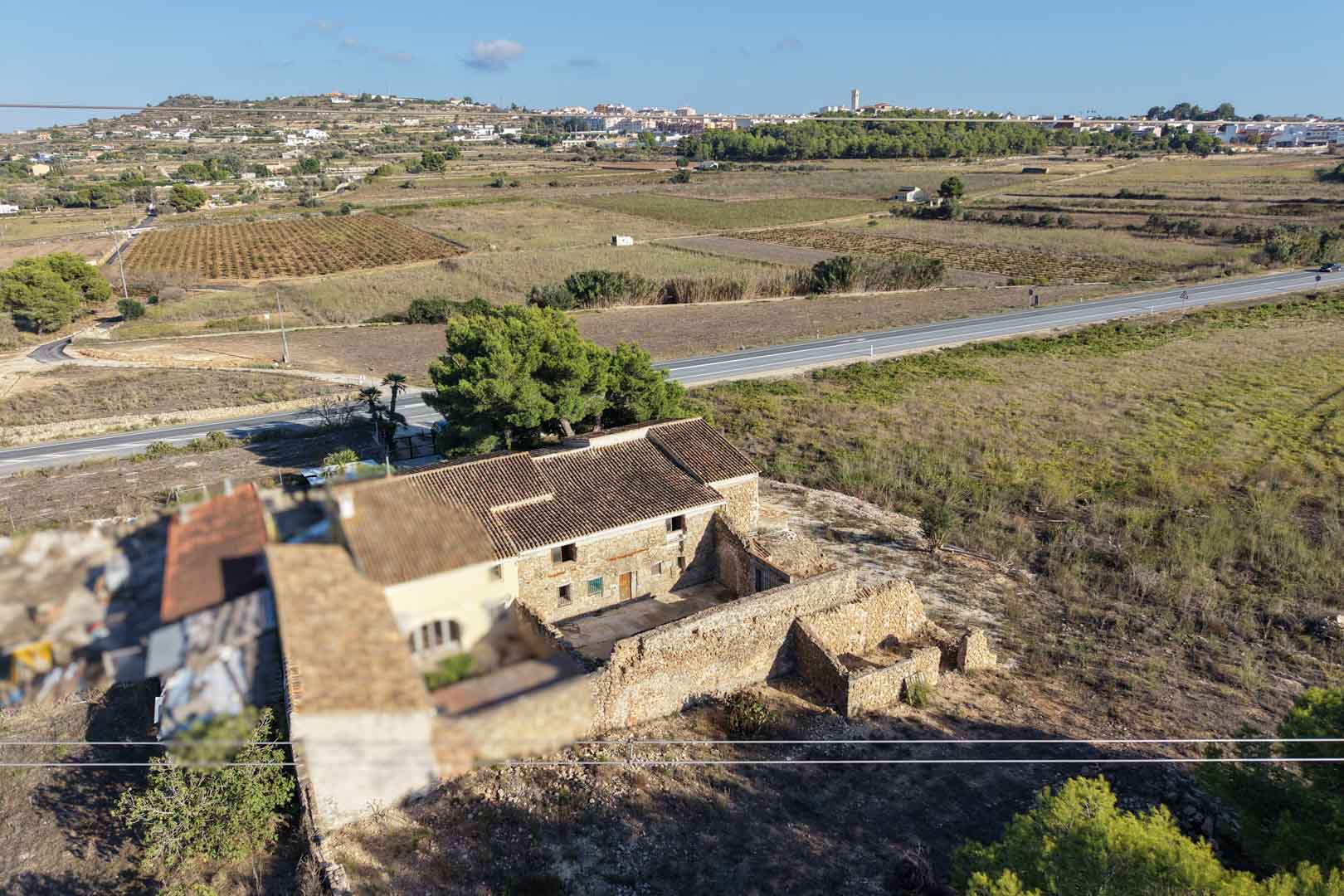 Vista aérea de la casa de piedra tradicional y sus anexos, situada en una amplia parcela rústica con vistas despejadas hacia el pueblo de Teulada.