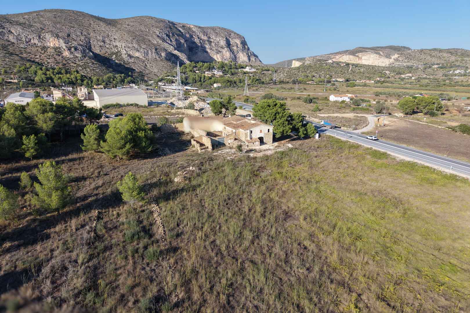 Vista aérea de la casa tradicional de piedra sobre una extensa parcela llana en Teulada, rodeada de vegetación mediterránea y con las montañas al fondo.