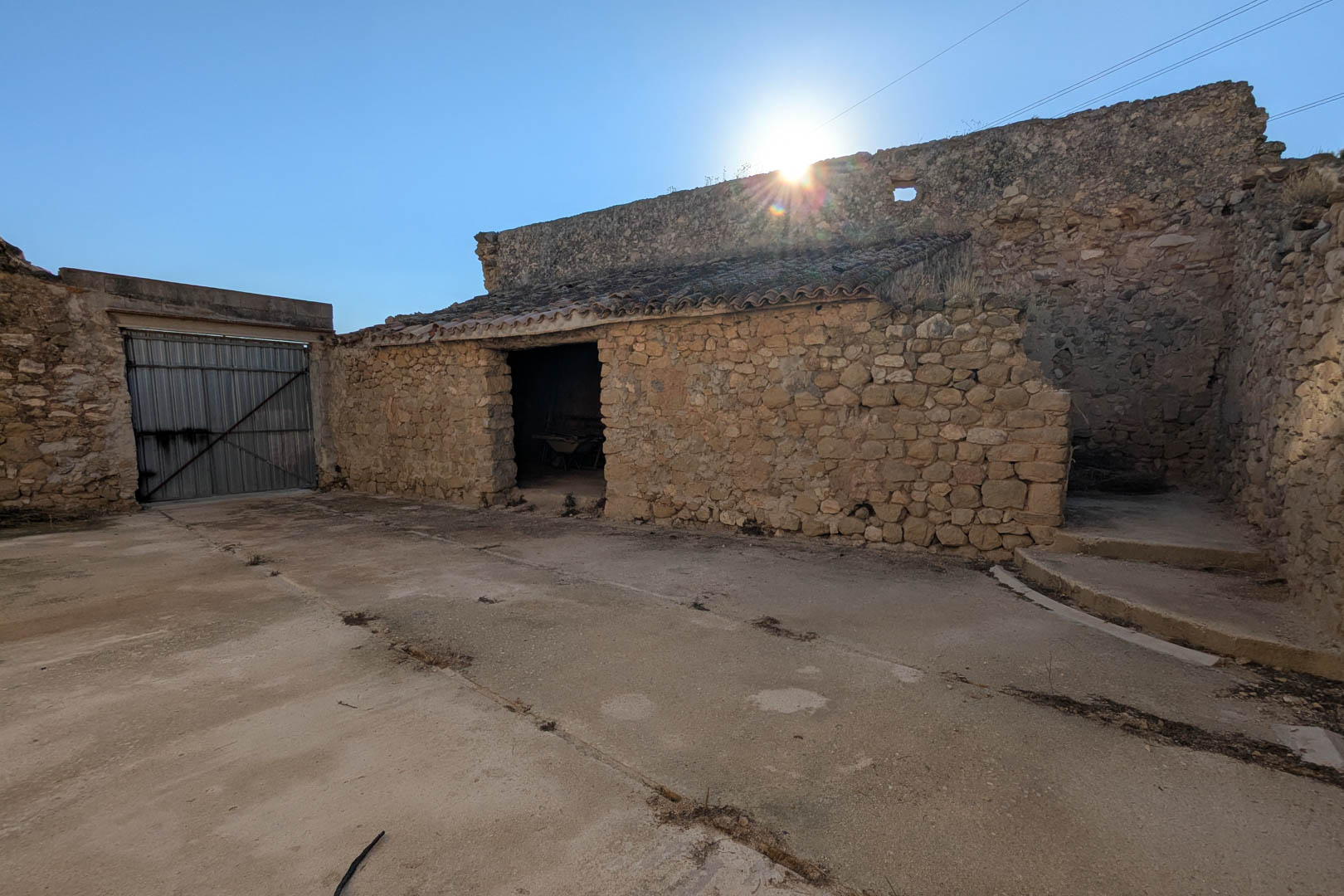 Vista exterior de los antiguos establos construidos en piedra natural, con techos de teja árabe y un amplio patio frontal de hormigón bajo el sol mediterráneo.