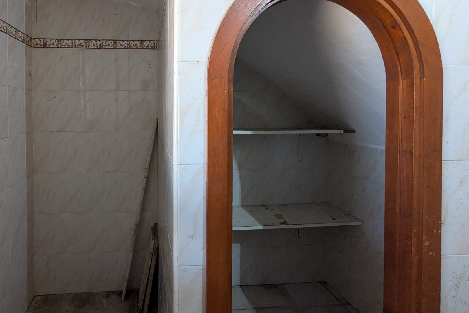 Zona de almacenaje bajo escalera con arco de madera, paredes alicatadas en blanco y estantes integrados en un espacio funcional.