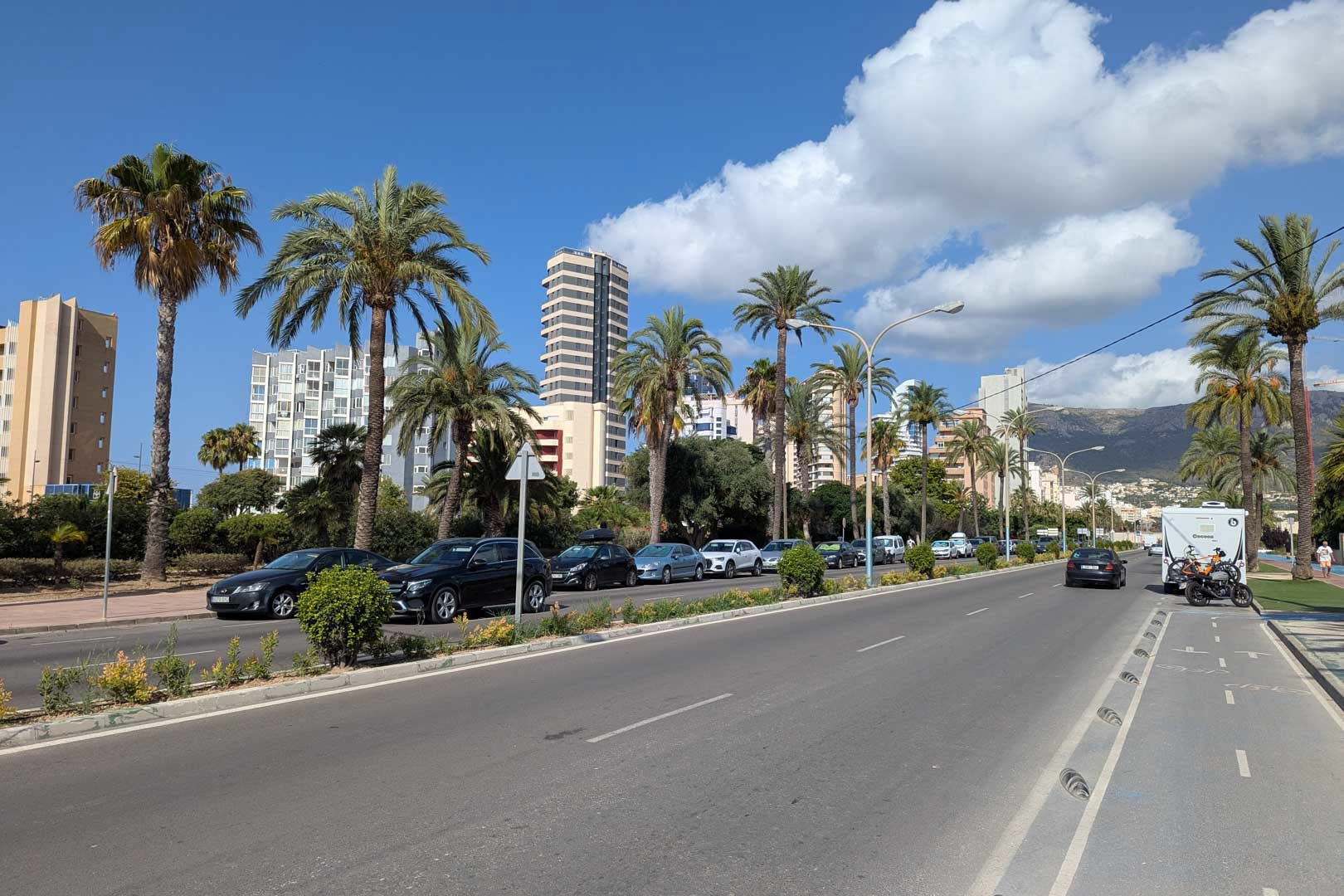 Avenida costera con carril bici, palmeras y edificios residenciales de gran altura bajo un cielo despejado con vistas a las montañas.