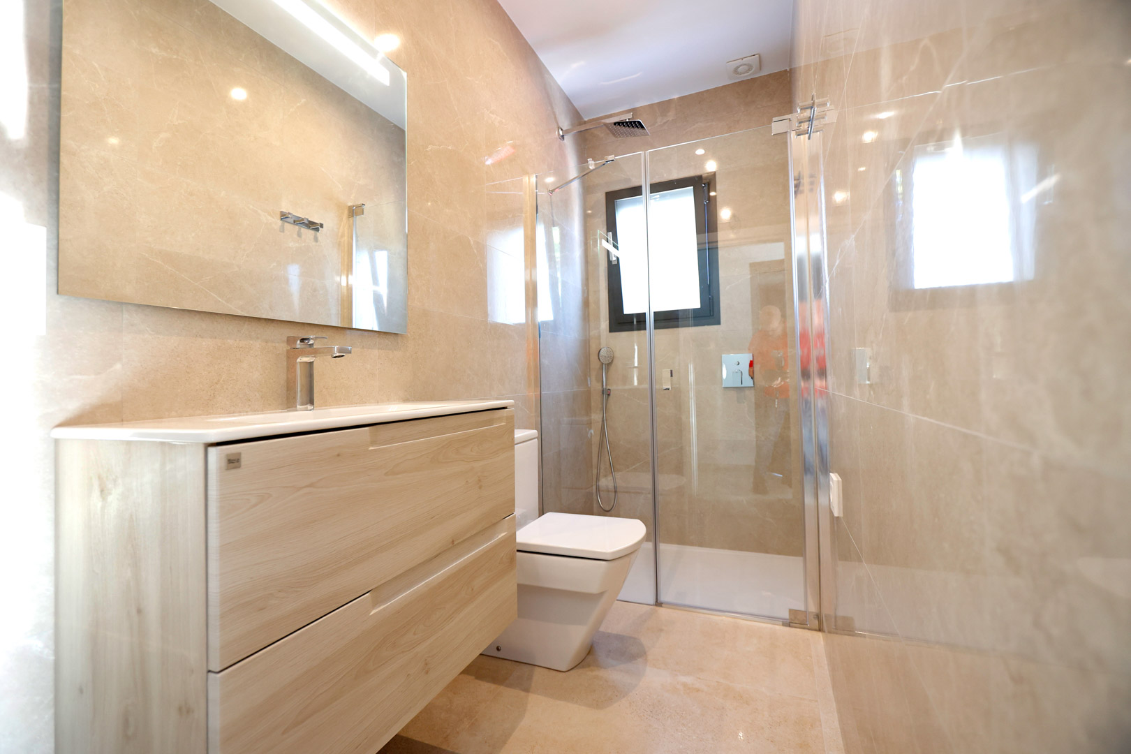 Baño contemporáneo con mueble de madera clara, ducha de cristal y paredes en gres porcelánico crema. Incluye grifería cromada e iluminación LED integrada.
