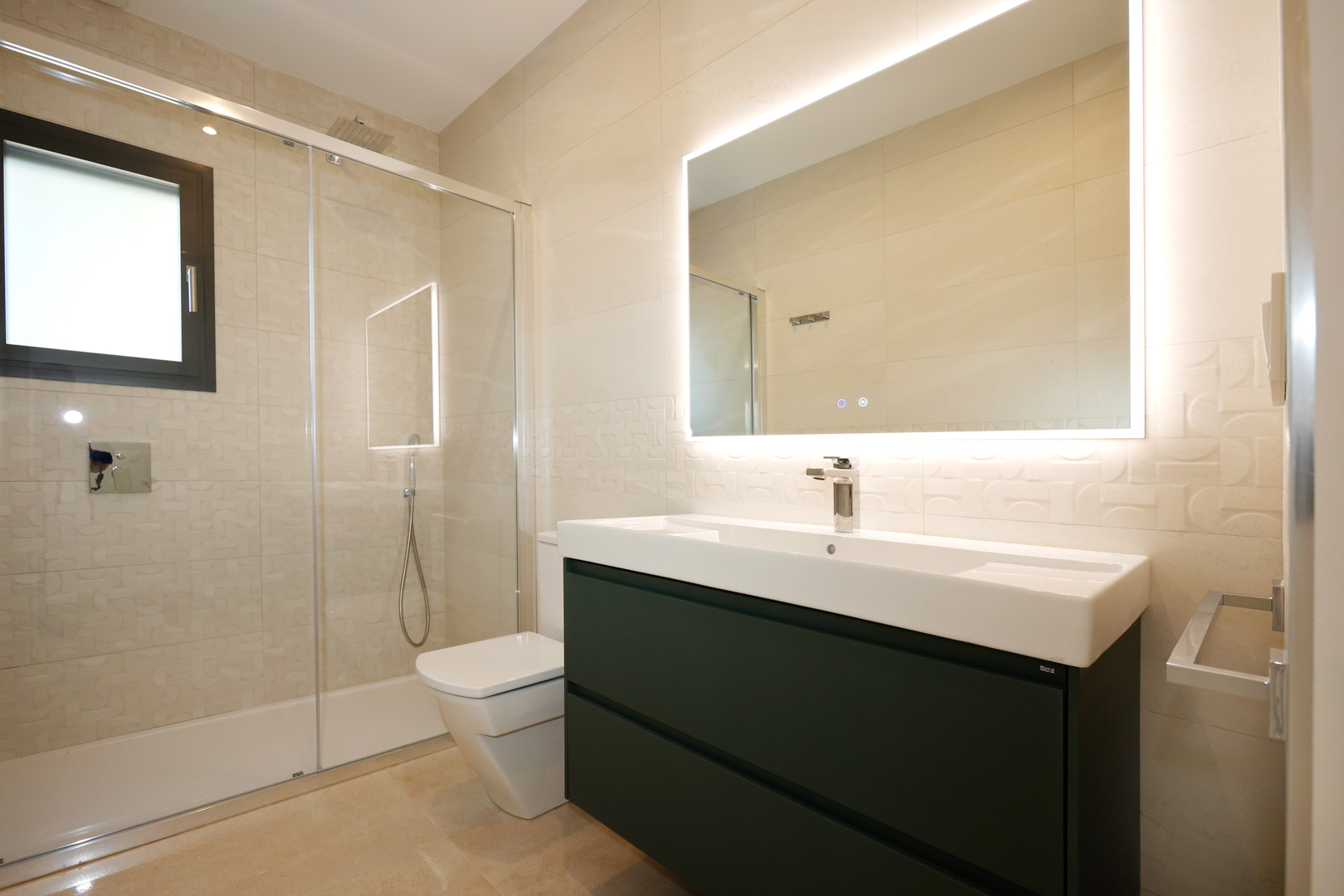 Baño contemporáneo con mueble suspendido antracita, lavabo Roca y espejo con iluminación LED. Dispone de ducha con mampara de cristal y paredes revestidas en gres porcelánico texturizado.