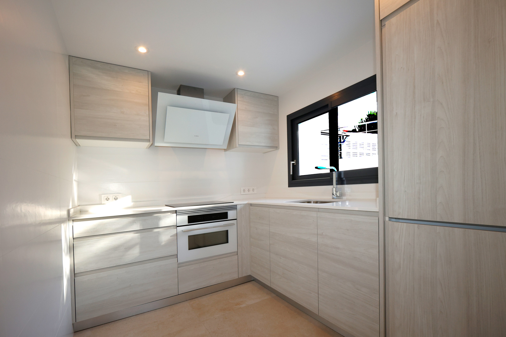 Cocina funcional con mobiliario de madera clara, encimera de Silestone blanco, placa de inducción, horno Bosch y campana extractora decorativa.