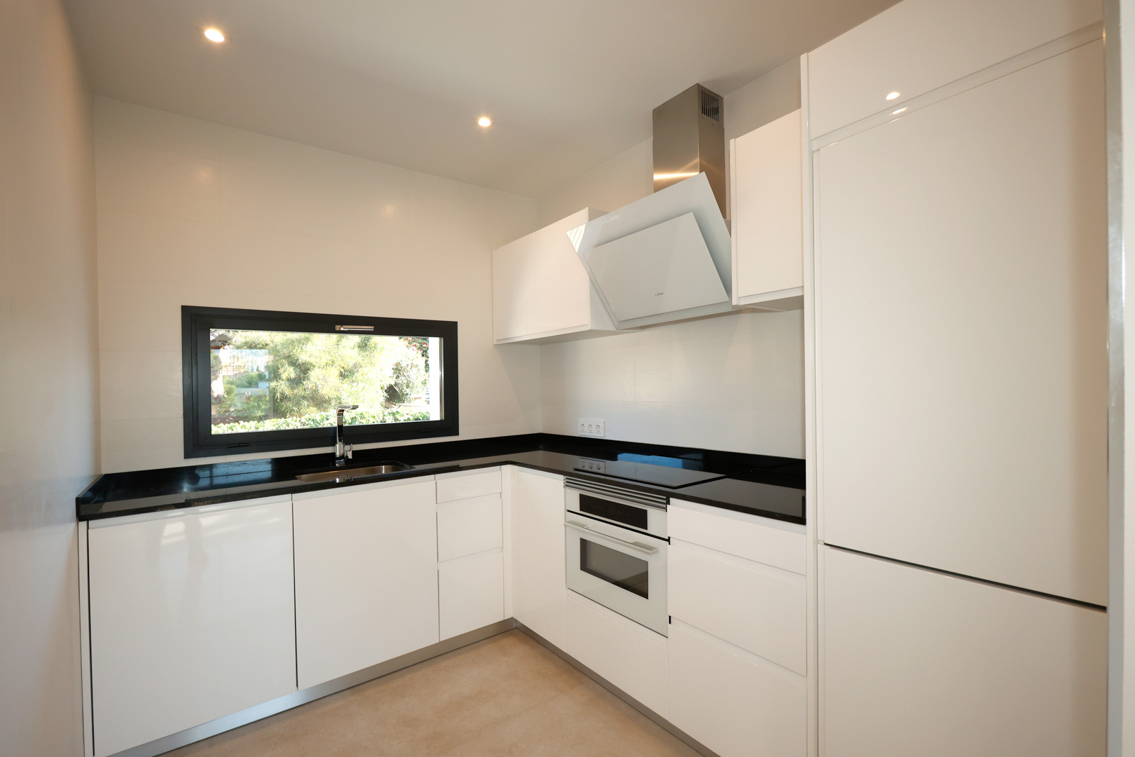 Cocina moderna con mobiliario blanco, encimera de Silestone negro, electrodomésticos Bosch integrados y ventana horizontal con vistas al exterior.