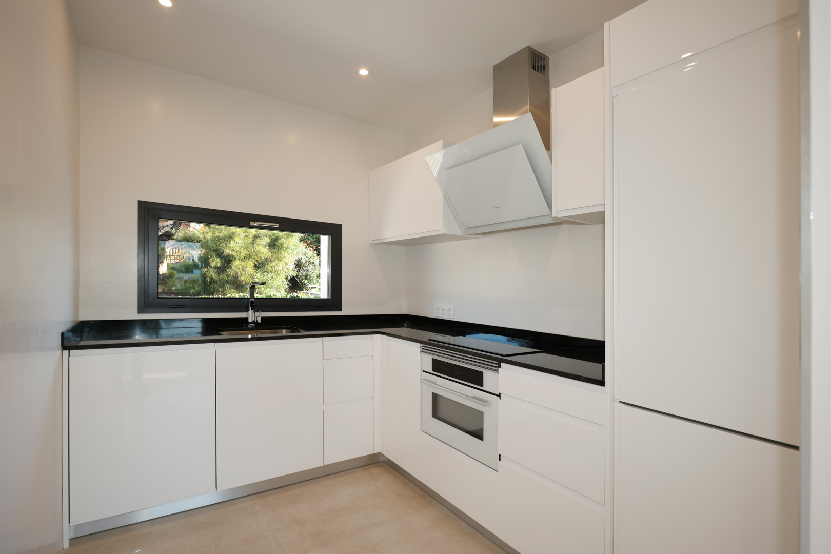 Cocina moderna con mobiliario blanco lacado, encimera de Silestone negro, electrodomésticos Bosch integrados y ventana panorámica hacia el jardín.