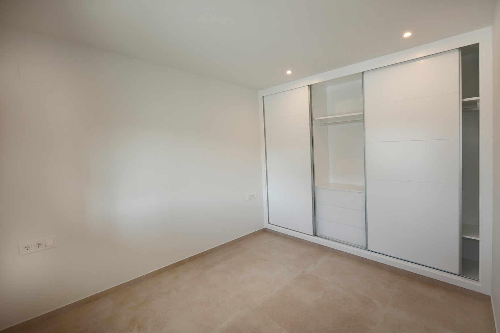 Dormitorio con armario empotrado de puertas correderas blancas lacadas, suelo de gres porcelánico y focos LED empotrados en el techo.