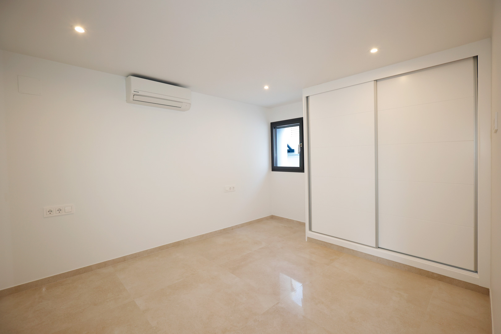 Dormitorio con armario empotrado de puertas correderas blancas, suelo de gres porcelánico, aire acondicionado y focos LED empotrados en el techo.