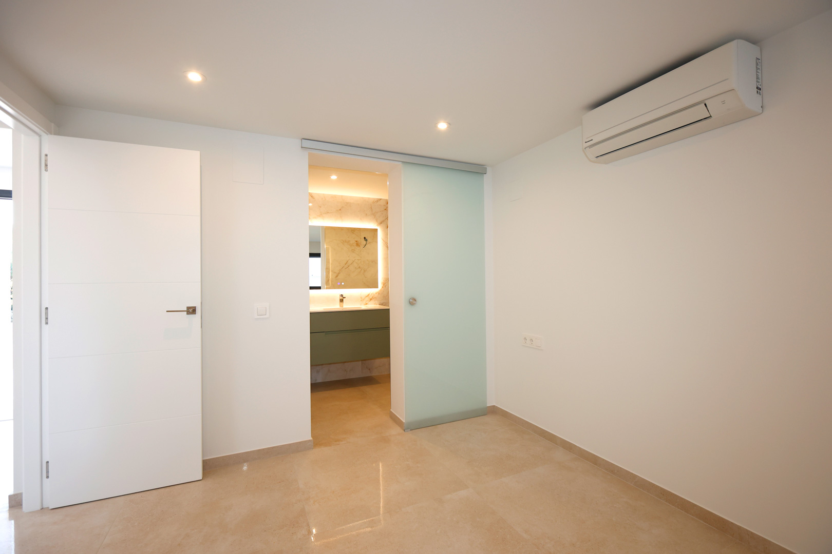 Dormitorio con suelo de gres porcelánico, aire acondicionado y acceso a baño en suite mediante puerta corredera de cristal translúcido.