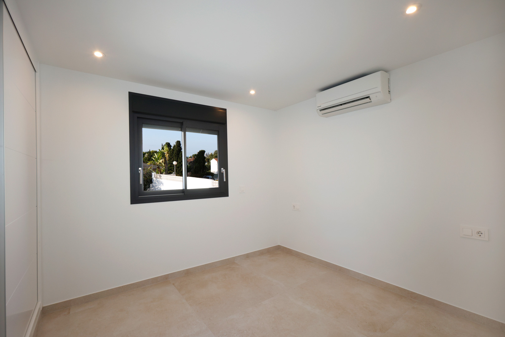 Dormitorio luminoso con suelo de gres porcelánico, iluminación LED empotrada, aire acondicionado y ventana de aluminio Technal con vistas al exterior.