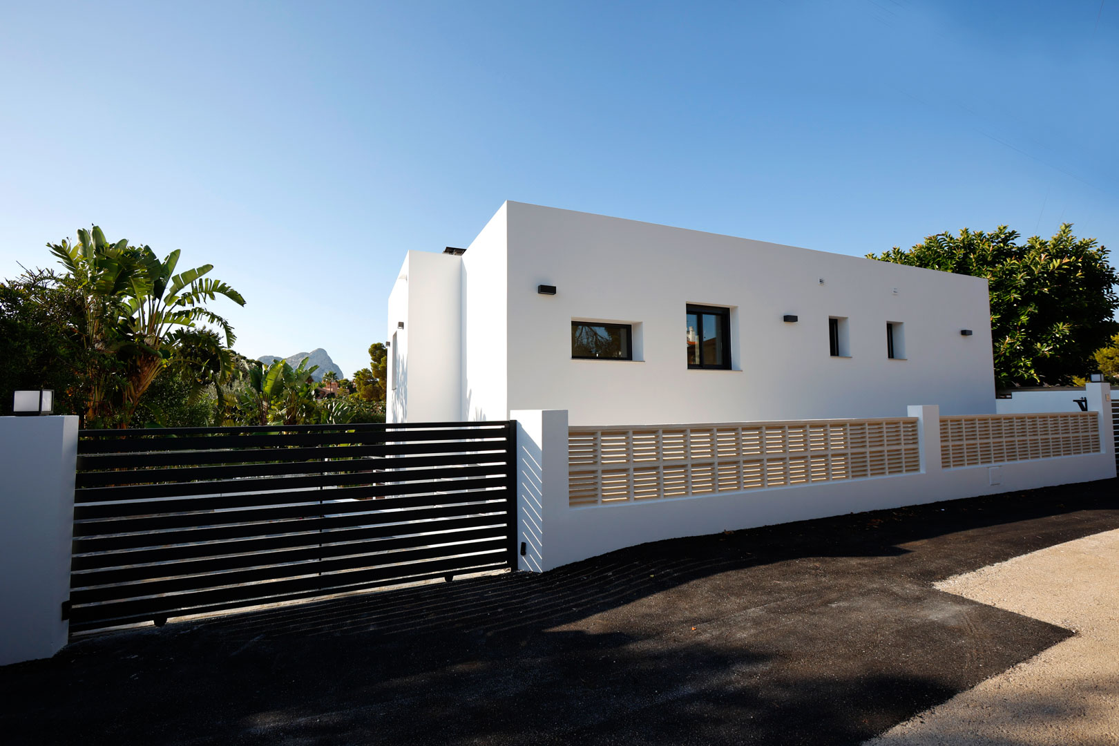 Fachada minimalista blanca con portón automático de lamas negras, muro perimetral con celosía y acceso pavimentado frente a vegetación mediterránea.