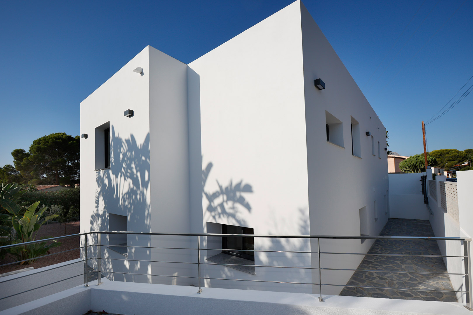 Fachada minimalista de líneas rectas con muros blancos, barandilla de acero inoxidable y suelo de piedra natural. Las sombras proyectadas resaltan el diseño contemporáneo.