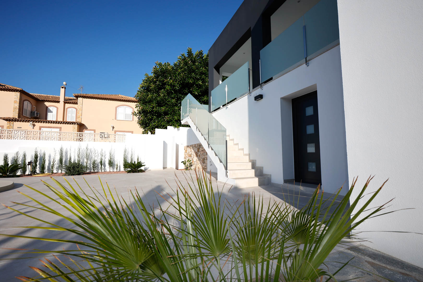 Fachada moderna con escalera exterior de gres, barandillas de cristal y acero, acceso independiente y terraza pavimentada con jardín perimetral.