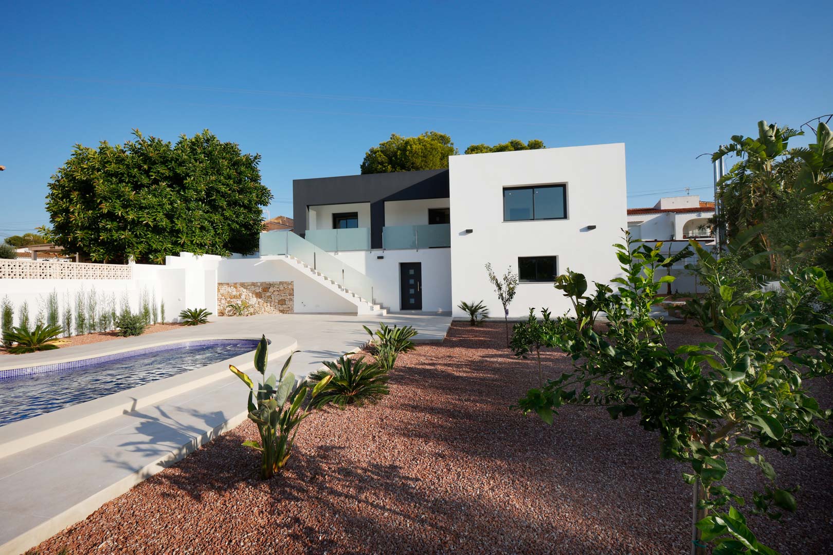 Fachada moderna con piscina, jardín de grava marmoleada roja y escalera exterior con barandillas de acero y cristal. Incluye muros de piedra natural y carpintería Technal.