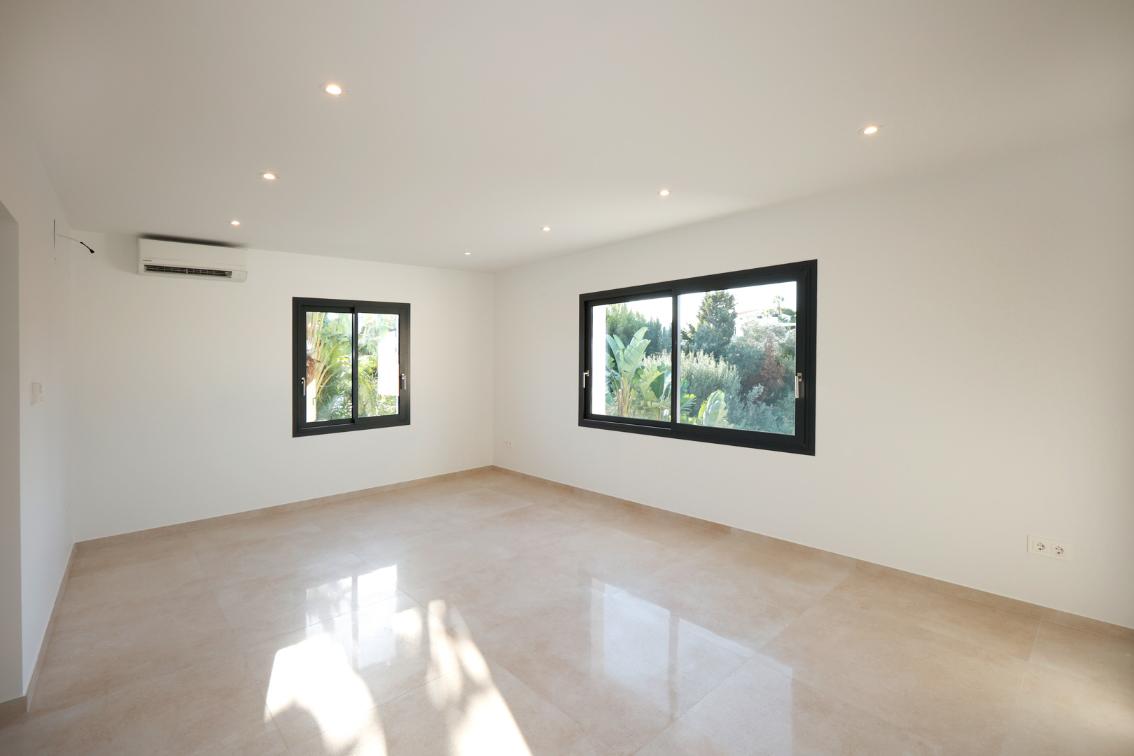 Habitación luminosa con suelo de gres porcelánico, iluminación LED integrada, aire acondicionado y ventanas de aluminio negro con vistas al jardín.