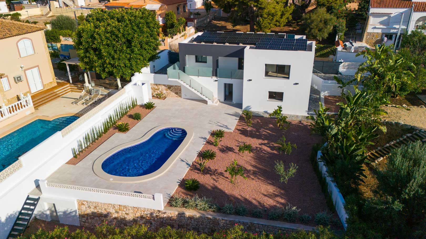 Vista aérea de la villa con piscina curva, jardín de grava roja y paneles solares en cubierta. Destacan las barandillas de cristal y muros de piedra natural.