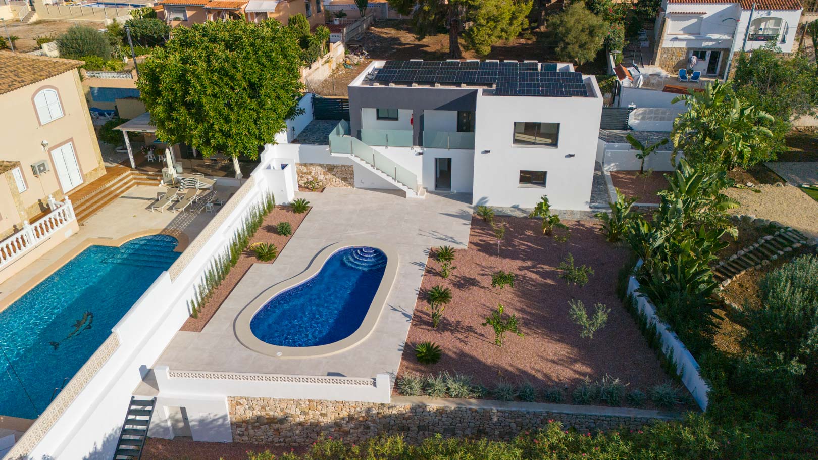 Vista aérea de la villa con piscina de diseño, jardín de grava roja y tejado con paneles solares. Destacan los muros de piedra natural y las barandillas de cristal en las terrazas.