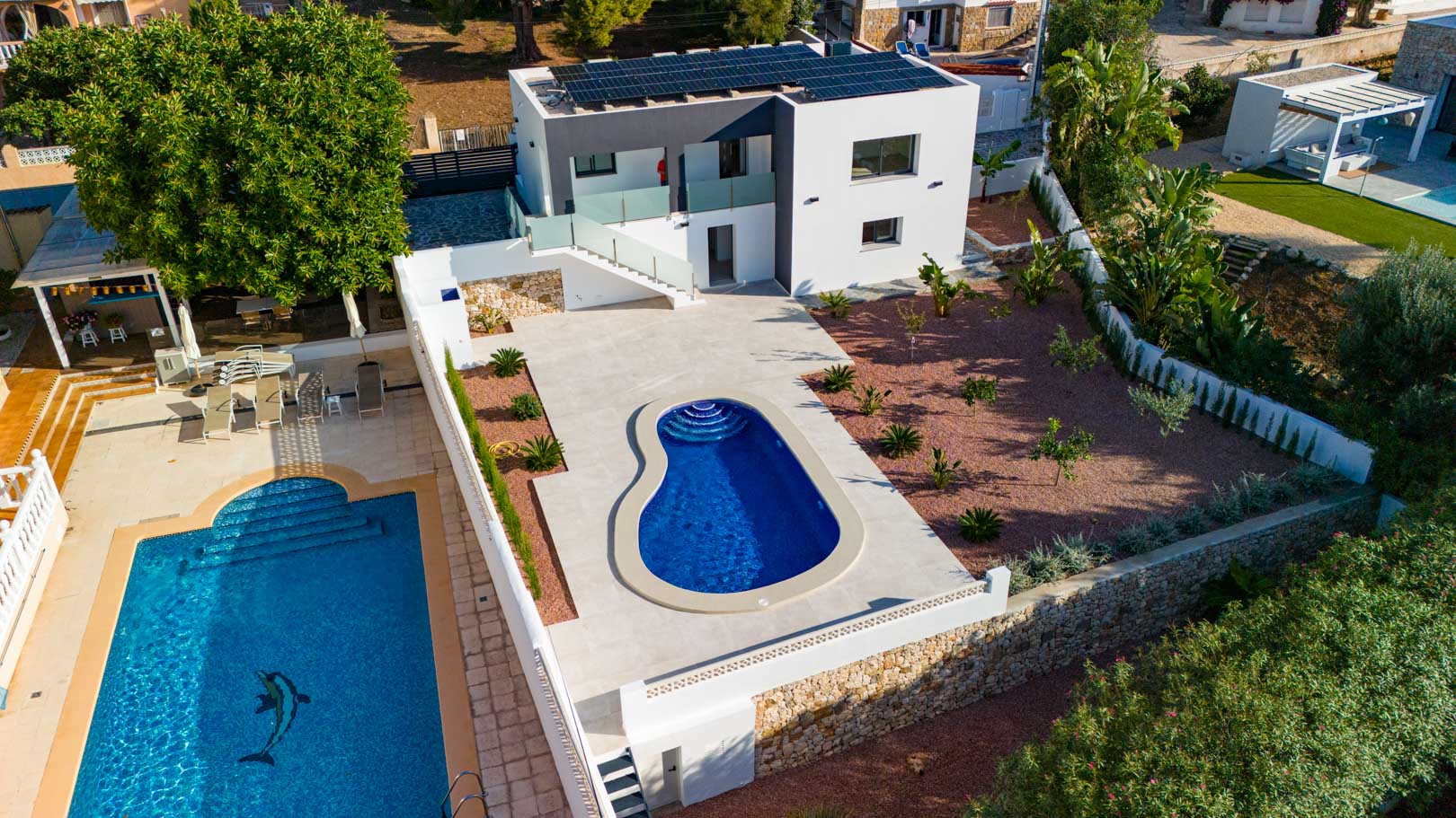 Vista aérea de la villa moderna con paneles solares, piscina de diseño orgánico, jardín de grava roja y muros de piedra natural. Las barandillas de cristal resaltan su estilo contemporáneo.