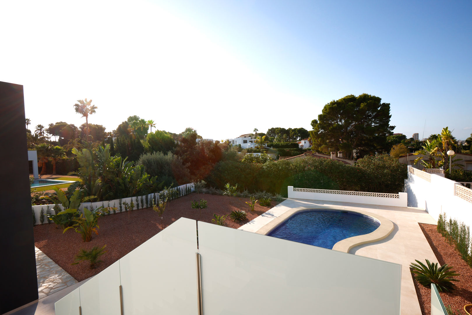 Vista desde la terraza superior con barandilla de cristal, mostrando la piscina curva, el jardín de grava marmoleada roja y el entorno mediterráneo.