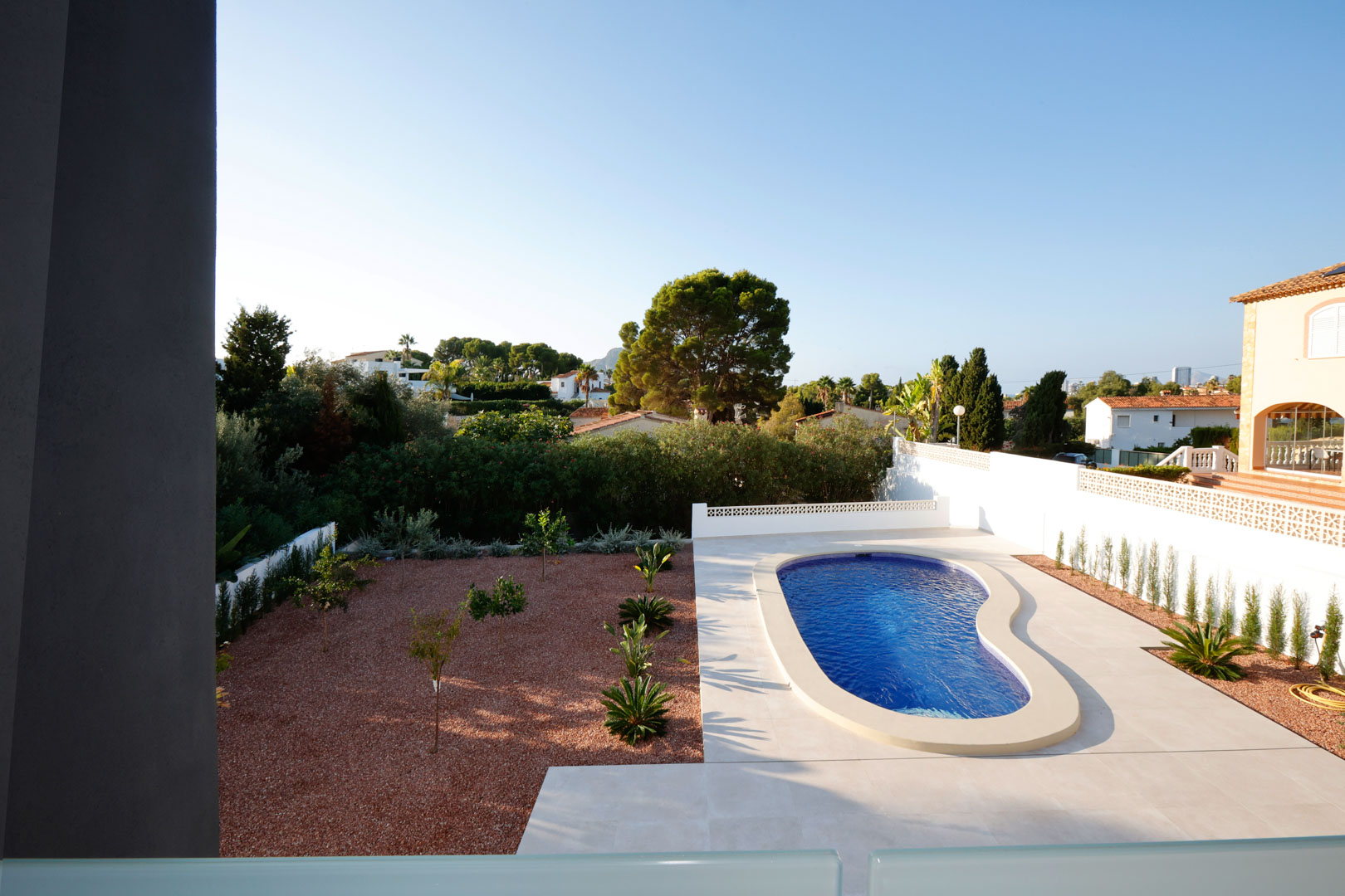 Vista elevada del jardín con piscina de diseño orgánico, rodeada de suelo porcelánico blanco y zona de grava marmoleada roja con vegetación mediterránea.