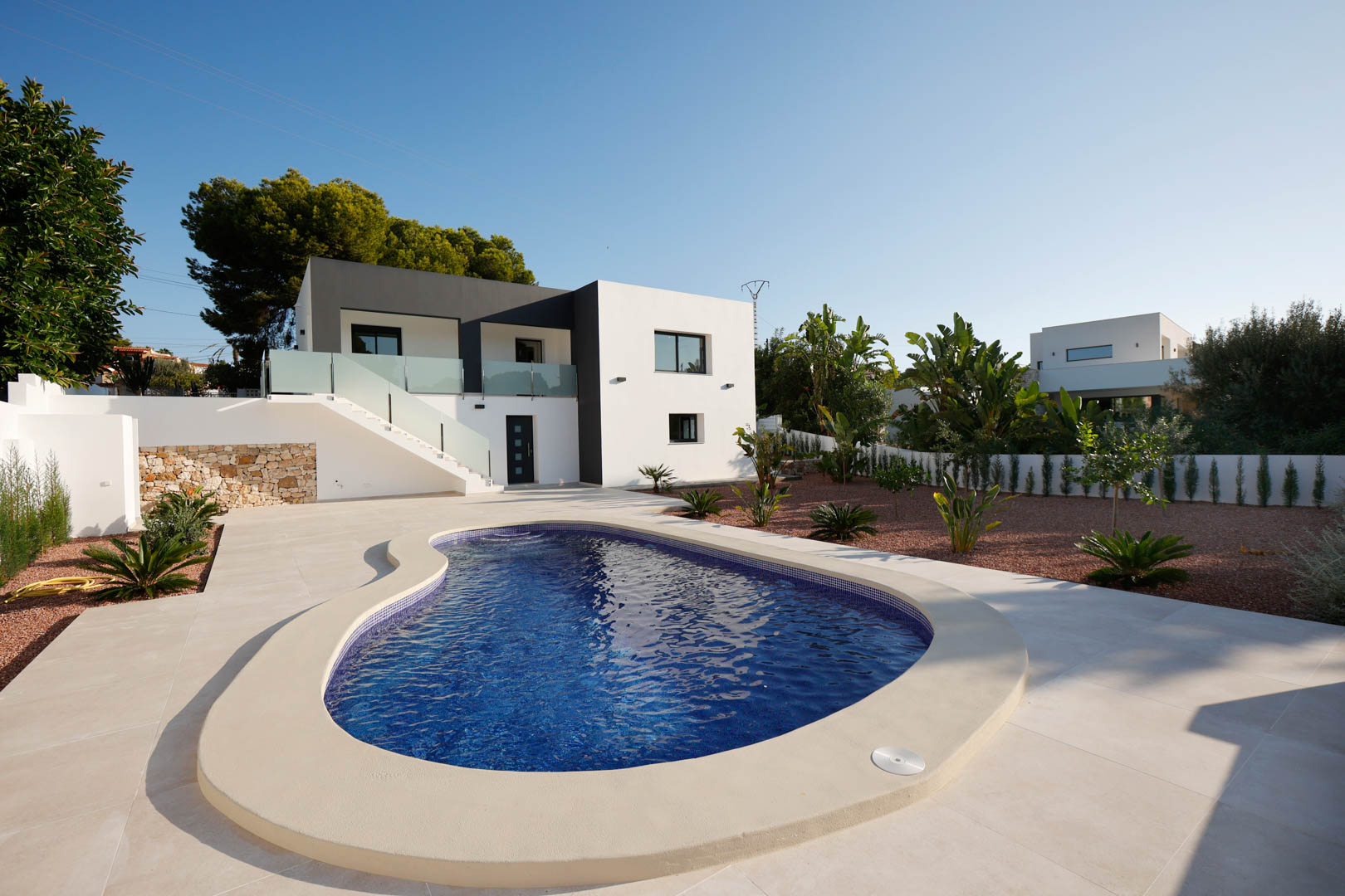 Vista exterior con piscina de diseño curvo, terraza de gres porcelánico y jardín de grava roja. Destaca la fachada bitono con escaleras y barandillas de cristal.