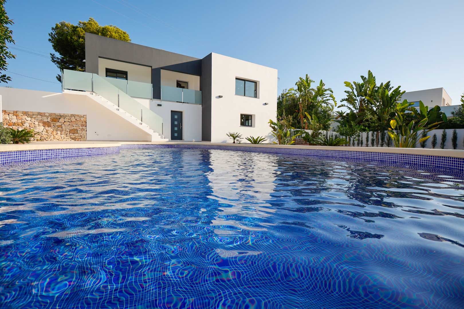 Vista exterior de la villa con piscina de gresite azul, fachada minimalista en blanco y gris, muros de piedra natural y terrazas con barandillas de cristal.