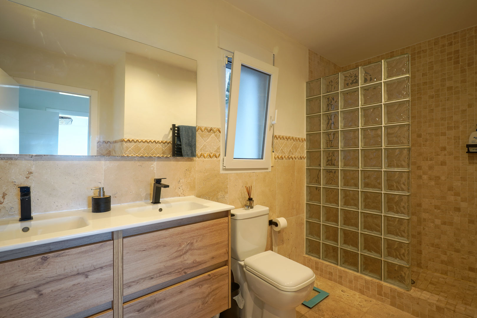 Baño moderno con mueble de doble lavabo en madera, grifería negra mate y ducha de obra con pared de pavés de cristal.