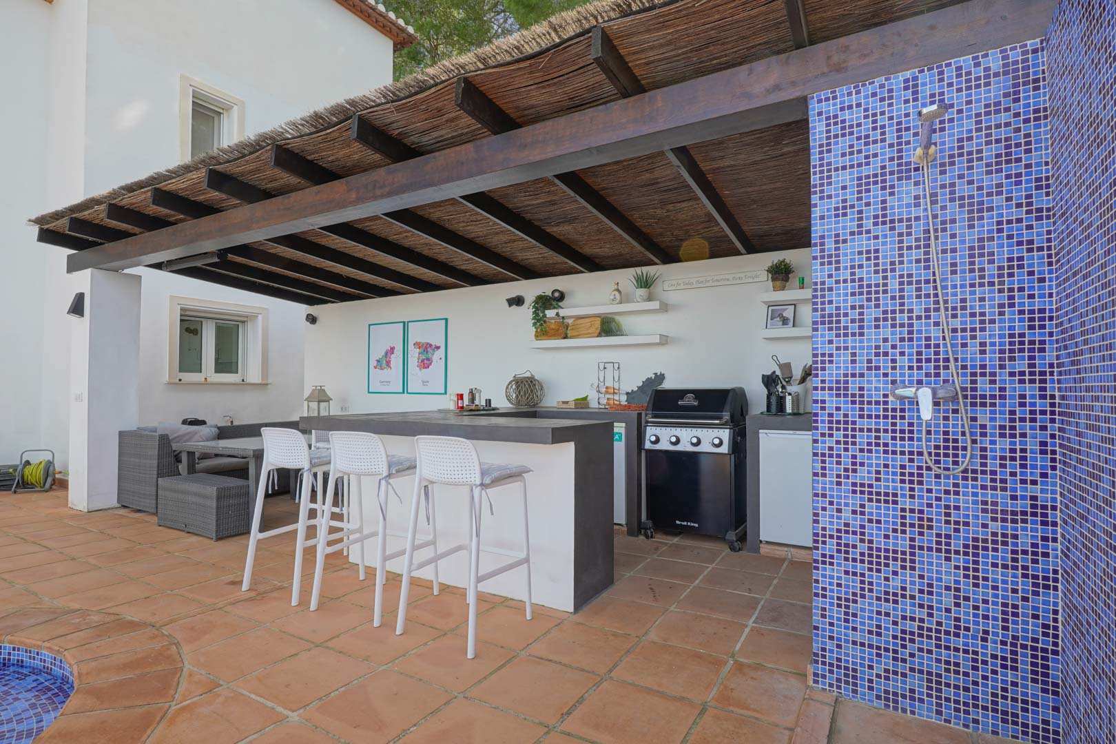 Cocina de verano exterior bajo pérgola de madera y brezo, equipada con barbacoa de gas, barra con taburetes y ducha exterior revestida en gresite azul.