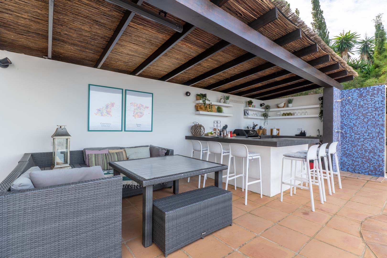 Cocina de verano y zona lounge bajo pérgola de vigas de madera y brezo, equipada con barra, taburetes blancos, mobiliario de ratán sintético y suelos de barro cocido.