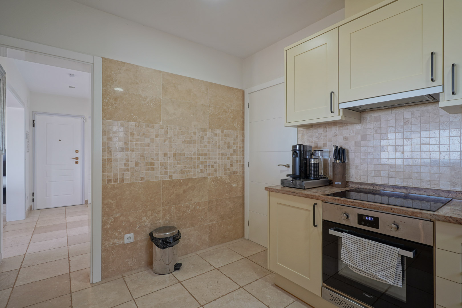 Cocina funcional con armarios crema, encimera de granito y electrodomésticos integrados. La pared luce un revestimiento de piedra natural con un elegante detalle de mosaico.